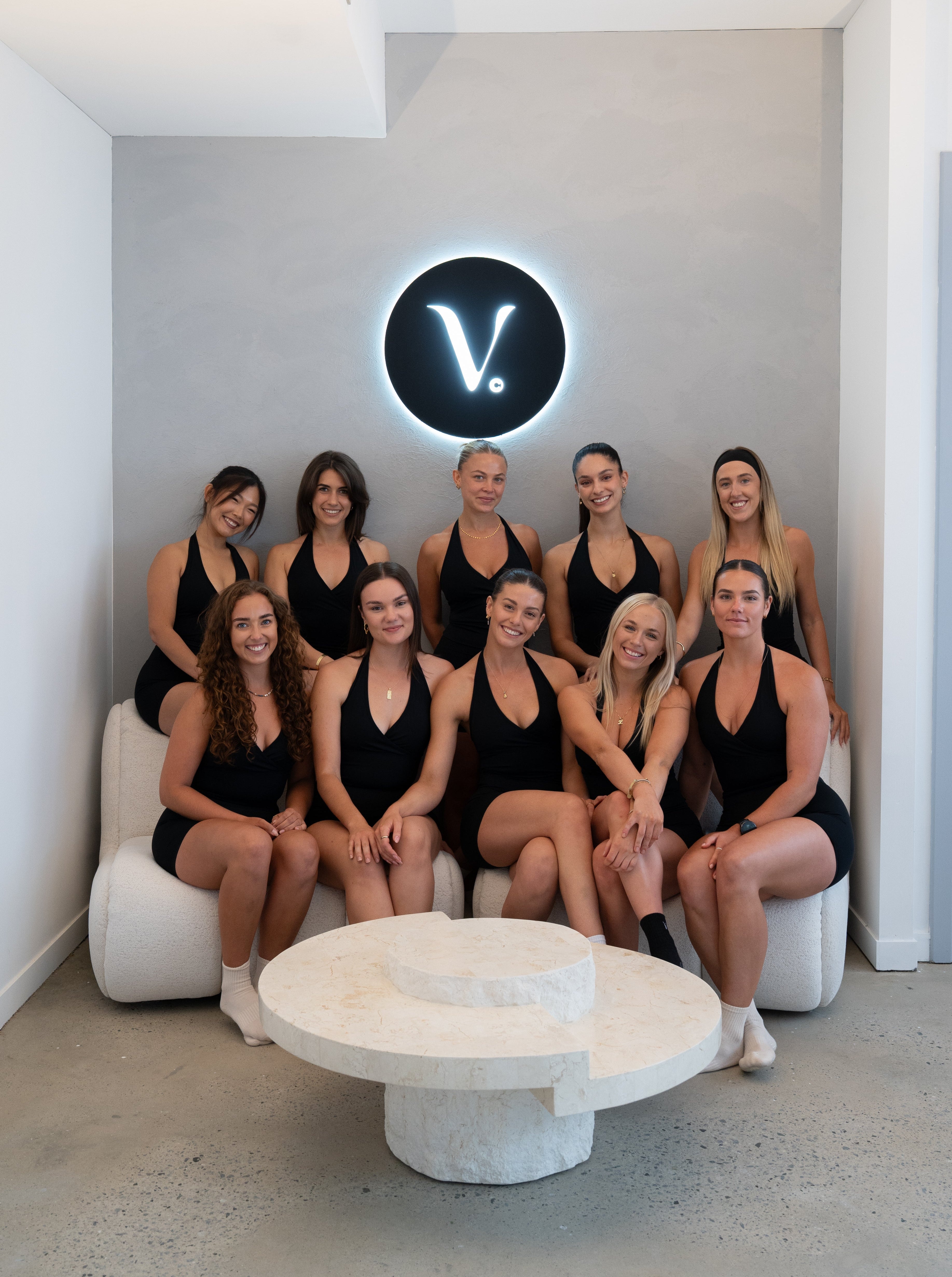 Group of women posing together in a modern interior setting with a 'V' logo on the wall.